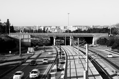 A highway with multiple lanes is visible, with several cars traveling in both directions. The road passes under an overpass, above which the skyline of a city with numerous buildings is visible. There are streetlights and a clear traffic separation, as well as some greenery in the form of trees on either side of the road.