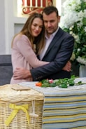 A couple embraces with solemn expressions in a setting that includes flowers and a wicker basket adorned with a yellow ribbon. The environment suggests a somber occasion, possibly a memorial or funeral.