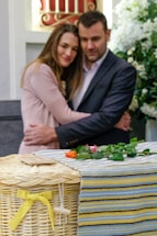 A couple embraces with solemn expressions in a setting that includes flowers and a wicker basket adorned with a yellow ribbon. The environment suggests a somber occasion, possibly a memorial or funeral.