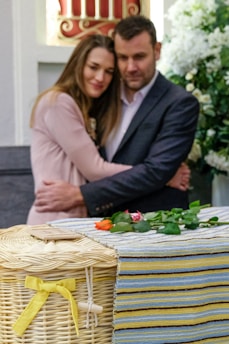 A couple embraces with solemn expressions in a setting that includes flowers and a wicker basket adorned with a yellow ribbon. The environment suggests a somber occasion, possibly a memorial or funeral.