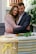A couple embraces with solemn expressions in a setting that includes flowers and a wicker basket adorned with a yellow ribbon. The environment suggests a somber occasion, possibly a memorial or funeral.
