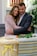 A couple embraces with solemn expressions in a setting that includes flowers and a wicker basket adorned with a yellow ribbon. The environment suggests a somber occasion, possibly a memorial or funeral.