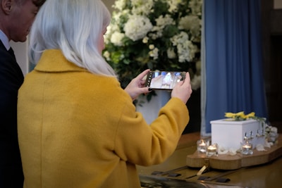 A person with long white hair wearing a yellow coat is taking a photo with a smartphone. There is a small white coffin on a wooden table adorned with flowers and lit candles in the foreground. Large white floral arrangements serve as the background decor.
