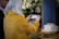 A person with long white hair wearing a yellow coat is taking a photo with a smartphone. There is a small white coffin on a wooden table adorned with flowers and lit candles in the foreground. Large white floral arrangements serve as the background decor.