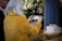 A person with long white hair wearing a yellow coat is taking a photo with a smartphone. There is a small white coffin on a wooden table adorned with flowers and lit candles in the foreground. Large white floral arrangements serve as the background decor.