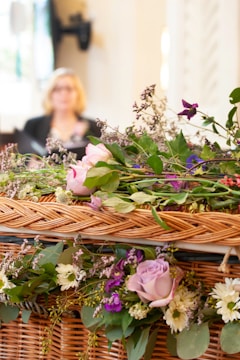 A wicker basket adorned with an array of flowers, including pink roses, purple and white blossoms, along with lush green leaves. In the background, a blurred figure of a person is visible.
