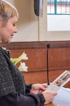 A person with short hair is reading from a card or booklet. The setting appears to be a formal or ceremonial event, possibly a speech or presentation, as indicated by the presence of a microphone on a stand. There are white lilies placed on a table in the background and a window with metal bars letting in natural light.