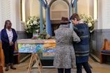 Inside a chapel, three individuals are near a colorfully painted coffin adorned with flowers. Two people stand close together, one wearing a leopard print coat and the other in a dark dress. The third person watches from a distance, wearing a navy blue cardigan and floral shirt. The space is decorated with bouquets of white flowers and pastel blue curtains.