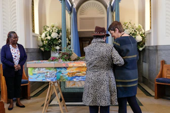 Inside a chapel, three individuals are near a colorfully painted coffin adorned with flowers. Two people stand close together, one wearing a leopard print coat and the other in a dark dress. The third person watches from a distance, wearing a navy blue cardigan and floral shirt. The space is decorated with bouquets of white flowers and pastel blue curtains.