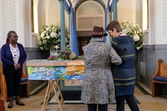Inside a chapel, three individuals are near a colorfully painted coffin adorned with flowers. Two people stand close together, one wearing a leopard print coat and the other in a dark dress. The third person watches from a distance, wearing a navy blue cardigan and floral shirt. The space is decorated with bouquets of white flowers and pastel blue curtains.