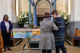 Inside a chapel, three individuals are near a colorfully painted coffin adorned with flowers. Two people stand close together, one wearing a leopard print coat and the other in a dark dress. The third person watches from a distance, wearing a navy blue cardigan and floral shirt. The space is decorated with bouquets of white flowers and pastel blue curtains.