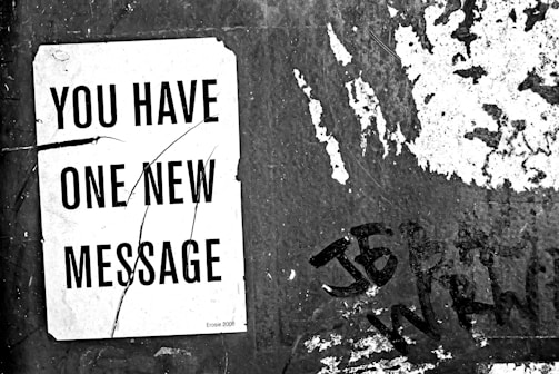 A black and white image containing a weathered wall with peeling paint and graffiti. Attached to the wall is a piece of white paper with large black text reading 'You Have One New Message'. The paper is worn and has a few creases.