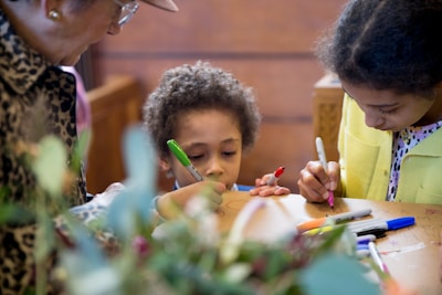 A group of children and an adult are engaged in a creative activity, drawing on a surface with colorful markers. The scene captures a warm and intimate moment of interaction and focus.