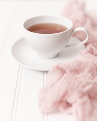 A white porcelain teacup filled with a light-colored tea sits on a matching saucer. Soft pink fabric gently drapes around the cup, adding a touch of color to the clean, white background. The overall composition exudes a sense of elegance and tranquility.