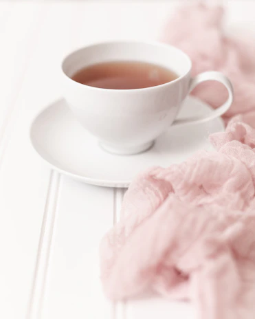 Elegant glass teacup on a beige linen cloth with subtle steam rising, surrounded by fresh green leaves.