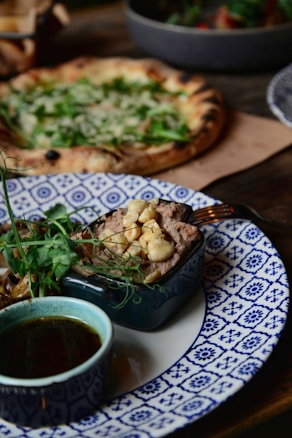 A beautifully plated dish features a small blue dish filled with what appears to be a savory pate topped with chickpeas, garnished with fresh herbs. This is placed on a patterned blue and white plate, alongside a small bowl of a dark dipping sauce. In the background, there is a rustic pizza topped with fresh greens.