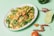 A vibrant dish of stir-fried mixed vegetables, including mushrooms, green bell peppers, carrots, and leafy greens, is served on a white plate. The plate is positioned on a pastel green background, surrounded by slices of lime, whole dried red chilies, a tomato, and other spices.