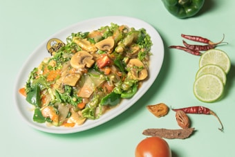 A vibrant dish of stir-fried mixed vegetables, including mushrooms, green bell peppers, carrots, and leafy greens, is served on a white plate. The plate is positioned on a pastel green background, surrounded by slices of lime, whole dried red chilies, a tomato, and other spices.