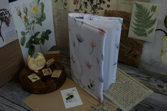 A table displays a closed book with a floral cover, surrounded by vintage botanical illustrations and handwritten letters. A wooden tray holds a moon-shaped candle and small vintage stamps. The scene exudes a sense of nostalgia and appreciation for nature.