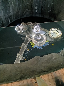 A unique underground space featuring a central platform with circular structures and luminous installations. The area includes a walkway bridge and several colorful boats arranged along the edge of a dark, expansive subterranean body of water. The surrounding walls and floor create a cavernous atmosphere.