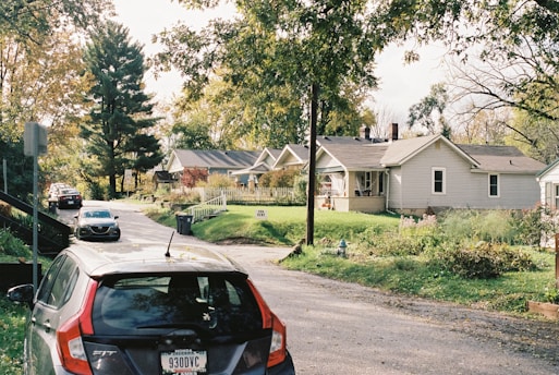 A peaceful suburban neighborhood with single-story homes lined up along a narrow street. The houses are surrounded by trees with lush green foliage. A few cars are parked along the street, which is bordered by a sidewalk. The sky is partly cloudy, and the overall atmosphere is calm and serene.