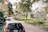 A peaceful suburban neighborhood with single-story homes lined up along a narrow street. The houses are surrounded by trees with lush green foliage. A few cars are parked along the street, which is bordered by a sidewalk. The sky is partly cloudy, and the overall atmosphere is calm and serene.