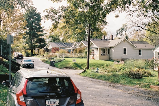 A peaceful suburban neighborhood with single-story homes lined up along a narrow street. The houses are surrounded by trees with lush green foliage. A few cars are parked along the street, which is bordered by a sidewalk. The sky is partly cloudy, and the overall atmosphere is calm and serene.