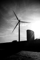 A large wind turbine stands silhouetted against a dramatic sky with wispy clouds and a bright sun. The scene is set in an industrial area with a cylindrical structure visible alongside the turbine.
