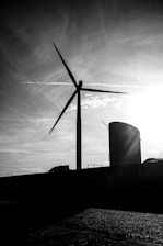 A large wind turbine stands silhouetted against a dramatic sky with wispy clouds and a bright sun. The scene is set in an industrial area with a cylindrical structure visible alongside the turbine.