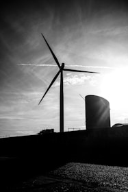 A large wind turbine stands silhouetted against a dramatic sky with wispy clouds and a bright sun. The scene is set in an industrial area with a cylindrical structure visible alongside the turbine.