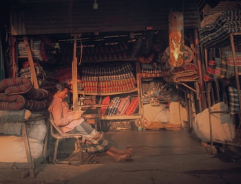 A person is sitting in a small shop that is filled with stacks of colorful, striped mattresses and fabrics. The person appears to be relaxing on a chair, checking something on a mobile phone. The shop has a cozy, slightly cluttered appearance with various textiles neatly arranged on shelves and hanging from the ceiling.
