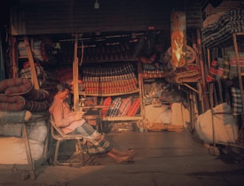 A person is sitting in a small shop that is filled with stacks of colorful, striped mattresses and fabrics. The person appears to be relaxing on a chair, checking something on a mobile phone. The shop has a cozy, slightly cluttered appearance with various textiles neatly arranged on shelves and hanging from the ceiling.
