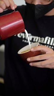 A person is pouring milk from a red pitcher into a brown cup. The individual is holding the cup with their left hand and appears to be wearing a black face mask and a black shirt.