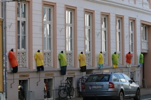 A row of mannequins dressed in colorful t-shirts and shorts is mounted on the exterior of a building with large windows. Below the mannequins, a bicycle and a parked car can be seen.