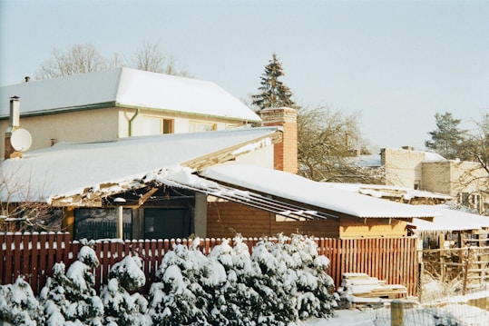 A residential area covered in snow, showing a house with a slanted roof, red brick chimney, and a wooden fence in the foreground. Snow-laden evergreen shrubs line the fence, while other houses are visible in the background amidst bare trees.