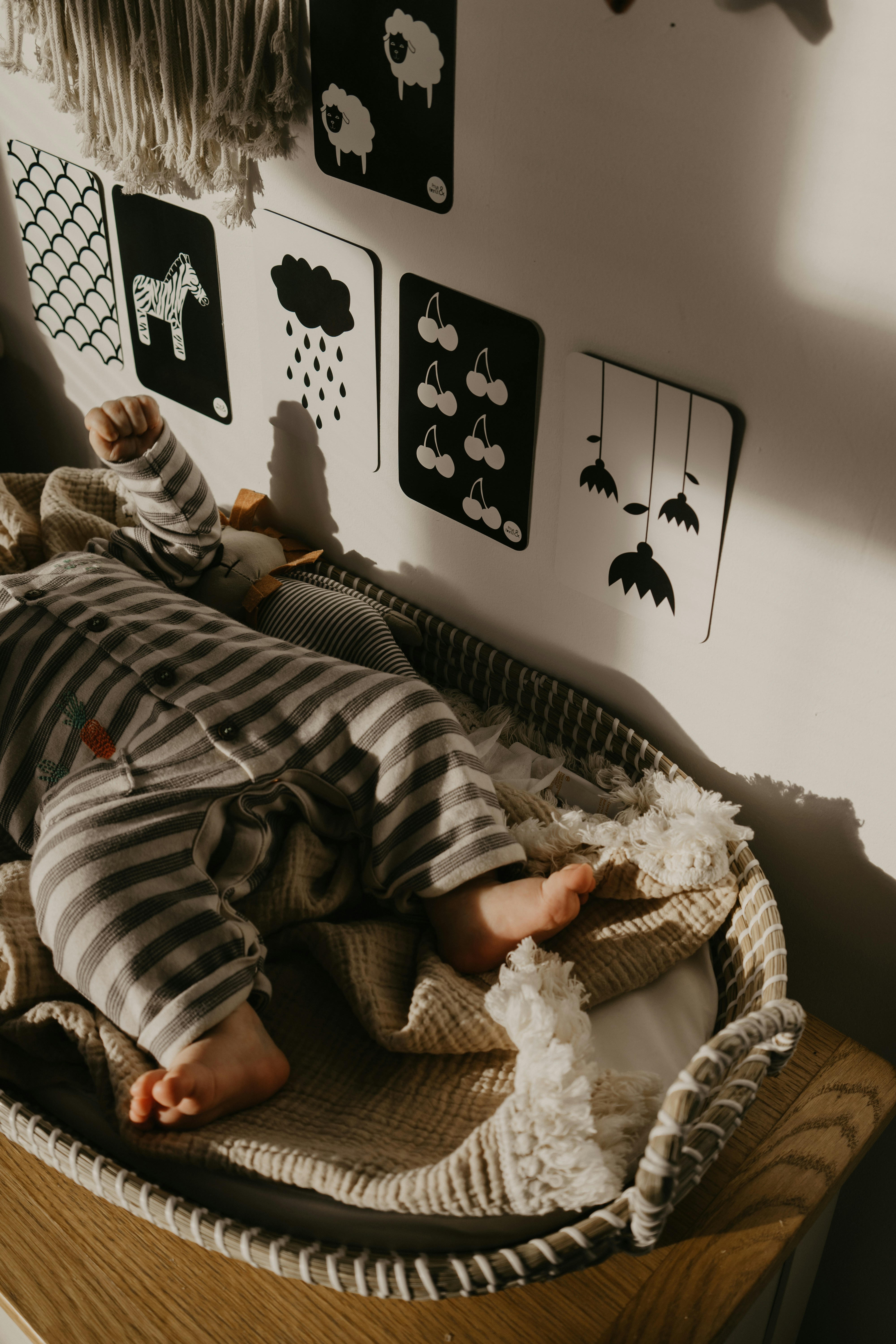 A baby dressed in striped pajamas sleeps peacefully in a woven bassinet. The wall behind features monochrome picture cards with simple black and white illustrations, including a zebra and raindrops. The soft light casts gentle shadows, adding a warm touch to the cozy setting.