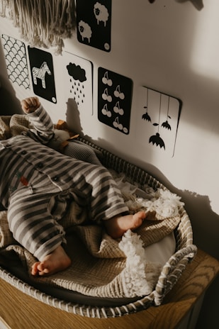 A baby dressed in striped pajamas sleeps peacefully in a woven bassinet. The wall behind features monochrome picture cards with simple black and white illustrations, including a zebra and raindrops. The soft light casts gentle shadows, adding a warm touch to the cozy setting.