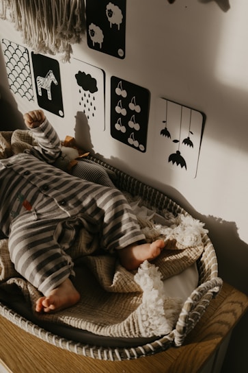 A baby dressed in striped pajamas sleeps peacefully in a woven bassinet. The wall behind features monochrome picture cards with simple black and white illustrations, including a zebra and raindrops. The soft light casts gentle shadows, adding a warm touch to the cozy setting.