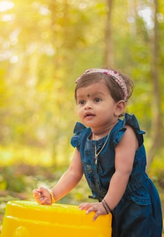 A toddler wearing a blue dress and a headband stands outdoors, surrounded by a lush, softly focused green background. The child is leaning on a bright yellow object, seemingly captivated by something out of view, while holding a small twig.