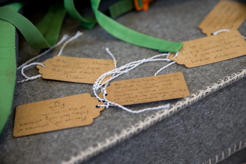 Several brown tags with handwritten notes are attached to white strings. The tags lie on a textured gray surface alongside green straps. The handwriting varies and appears personal, conveying individual messages.