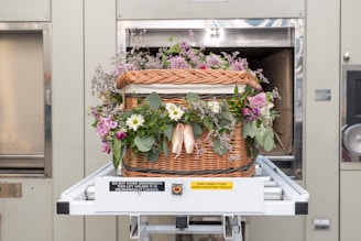 A woven wicker basket adorned with a variety of colorful flowers is centered on a mechanical trolley system inside a facility. The basket includes pink roses, white daisies, and lush green foliage, and is strategically placed in front of a large metallic door. There are warning labels on the trolley, emphasizing safety precautions.