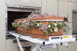A wicker casket adorned with a variety of flowers, including purple roses and green foliage, is being placed inside a cremation chamber. The casket rests on a metal trolley, and the surroundings are industrial with metallic tones.