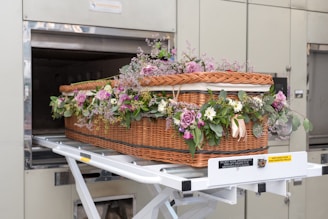 A wicker casket adorned with a variety of flowers, including purple roses and green foliage, is being placed inside a cremation chamber. The casket rests on a metal trolley, and the surroundings are industrial with metallic tones.