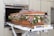 A wicker casket adorned with a variety of flowers, including purple roses and green foliage, is being placed inside a cremation chamber. The casket rests on a metal trolley, and the surroundings are industrial with metallic tones.