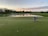 A golfer is crouched on the green, carefully lining up a putt with a golf ball nearby. The scene takes place during sunset, reflecting off a serene water hazard surrounded by lush greenery and trees. The sky is partly cloudy with hues of orange and blue.