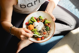 A person is seated with a colorful, healthy bowl of food on their lap, containing quinoa, broccoli, cherry tomatoes, and dollops of sauce. The person is using a fork and wearing a light-colored sleeveless top with a visible text print.