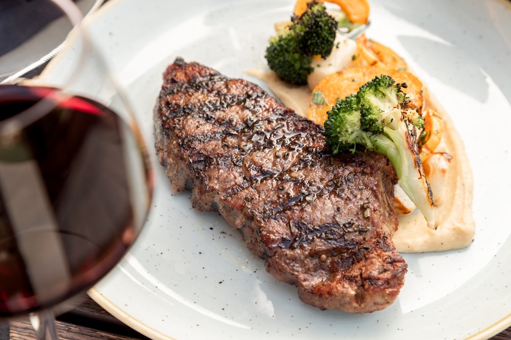 a white plate topped with meat and veggies next to a glass of wine