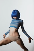 A person wearing a blue wig and a sheer blue striped top paired with beige shorts. The pose is dynamic, and the head is tilted downward, obscuring part of the face.