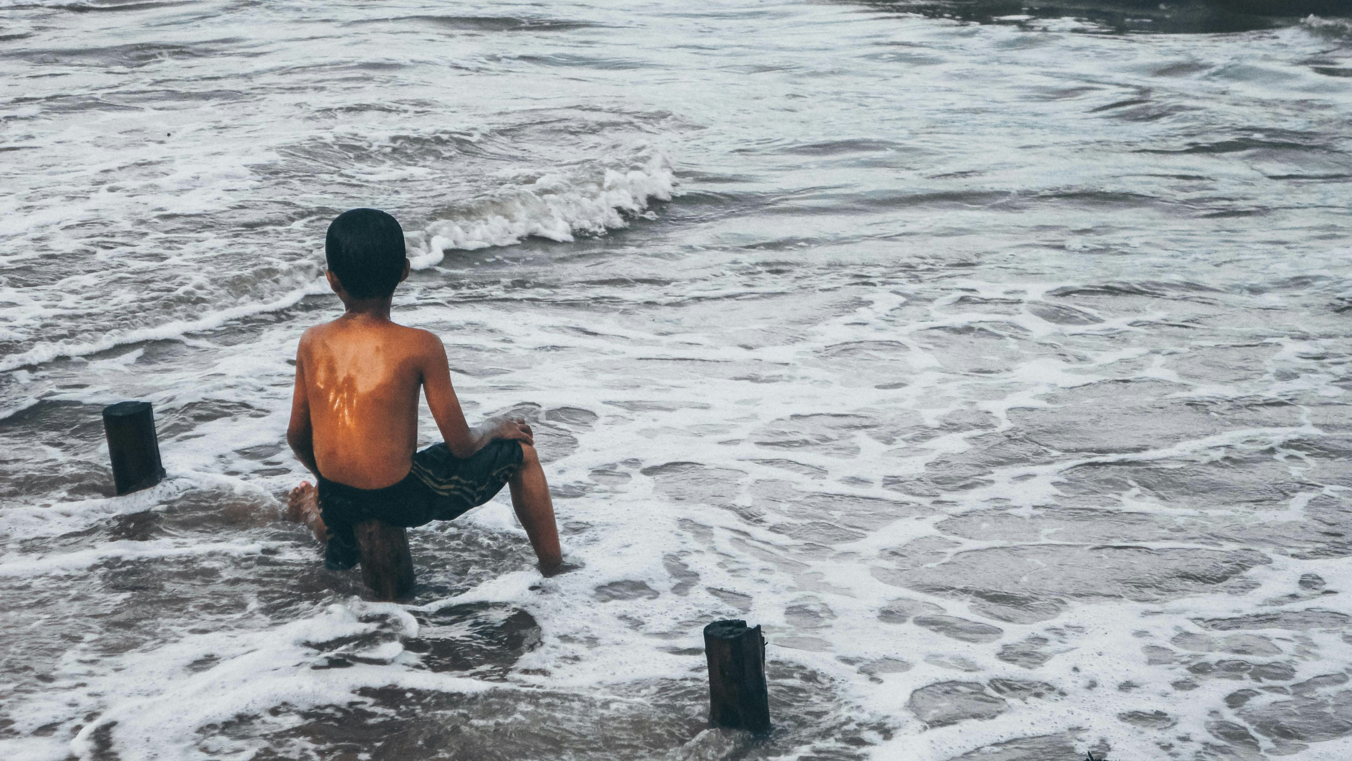 A boy sits on a post, facing the sea.