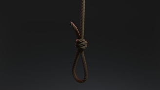 A tightly knotted rope hangs vertically against a dark, shadowy background. The rope's fibers are clearly visible, emphasizing its rough texture.